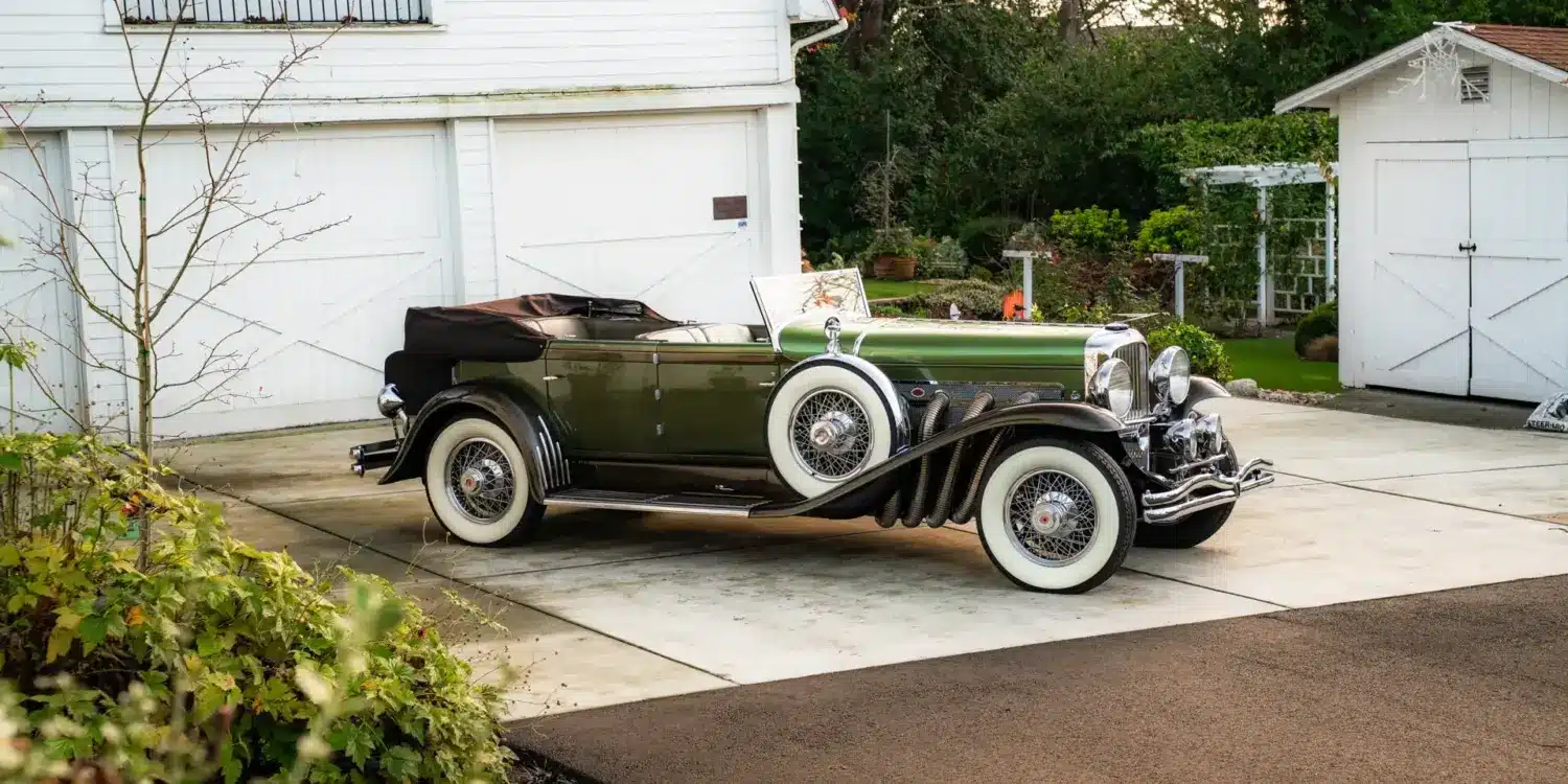 Duesenberg Model SJ Cabriolet-Sedan Baujahr 1929, grün lackiert, mit Verdeck, seitlich vor einer Garage geparkt.