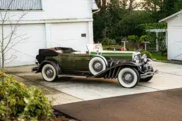 Duesenberg Model SJ Cabriolet-Sedan Baujahr 1929, grün lackiert, mit Verdeck, seitlich vor einer Garage geparkt.