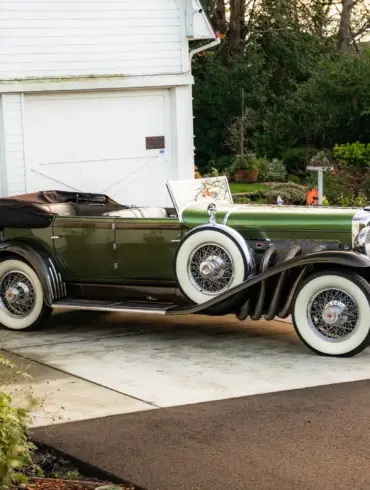 Duesenberg Model SJ Cabriolet-Sedan Baujahr 1929, grün lackiert, mit Verdeck, seitlich vor einer Garage geparkt.