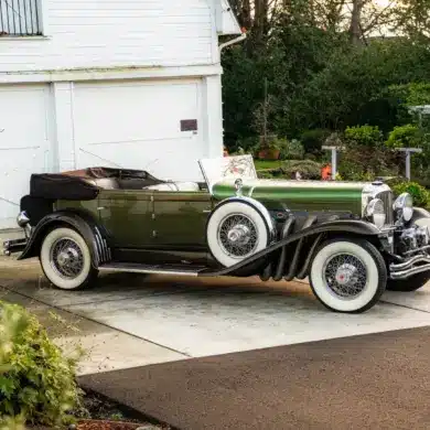 Duesenberg Model SJ Cabriolet-Sedan Baujahr 1929, grün lackiert, mit Verdeck, seitlich vor einer Garage geparkt.
