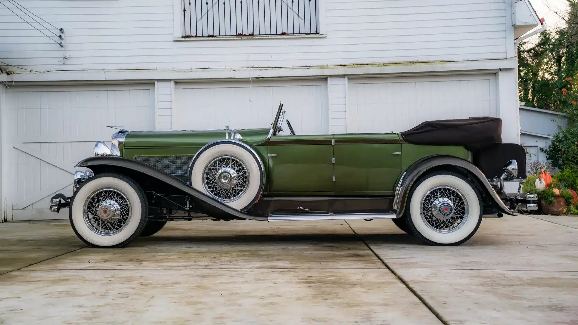 Seitliche Ansicht eines zweifarbig grünen Duesenberg Model SJ Cabriolet-Sedan von 1929 mit weißwandbereiften Speichenrädern, aufgenommen vor einer weißen Garage.