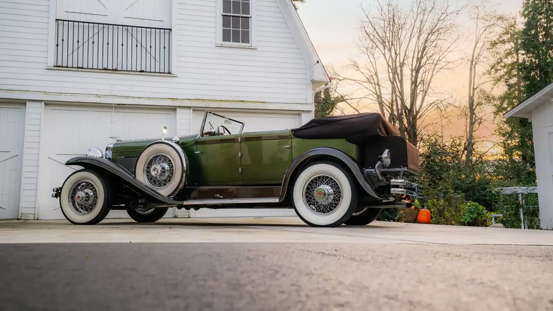 Restauriertes Duesenberg Model SJ Cabriolet-Sedan von 1929 mit grüner Lackierung und weißwandreifen vor einer weißen Garage bei Sonnenuntergang.