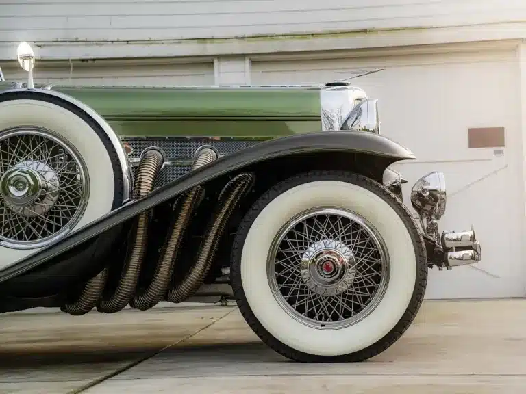 Detailaufnahme der Vorderachse eines grünen Duesenberg Model SJ Cabriolet-Sedan von 1929, mit chromglänzenden Speichenrädern, weißen Reifen und markanten seitlichen Auspuffrohren.