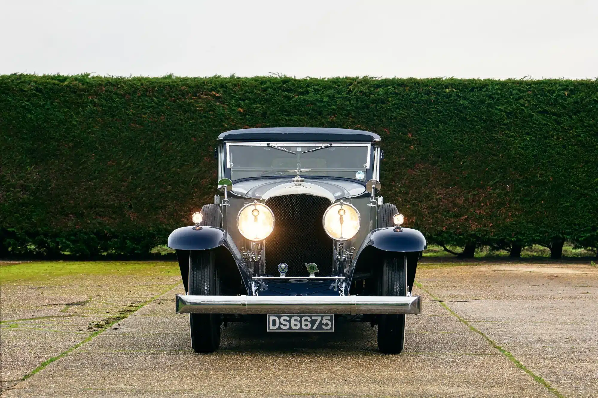 Frontansicht eines Bentley Speed Six von 1930 mit Schutte-Karosserie, Scheinwerfer eingeschaltet, imposante Erscheinung vor einer grünen Hecke.