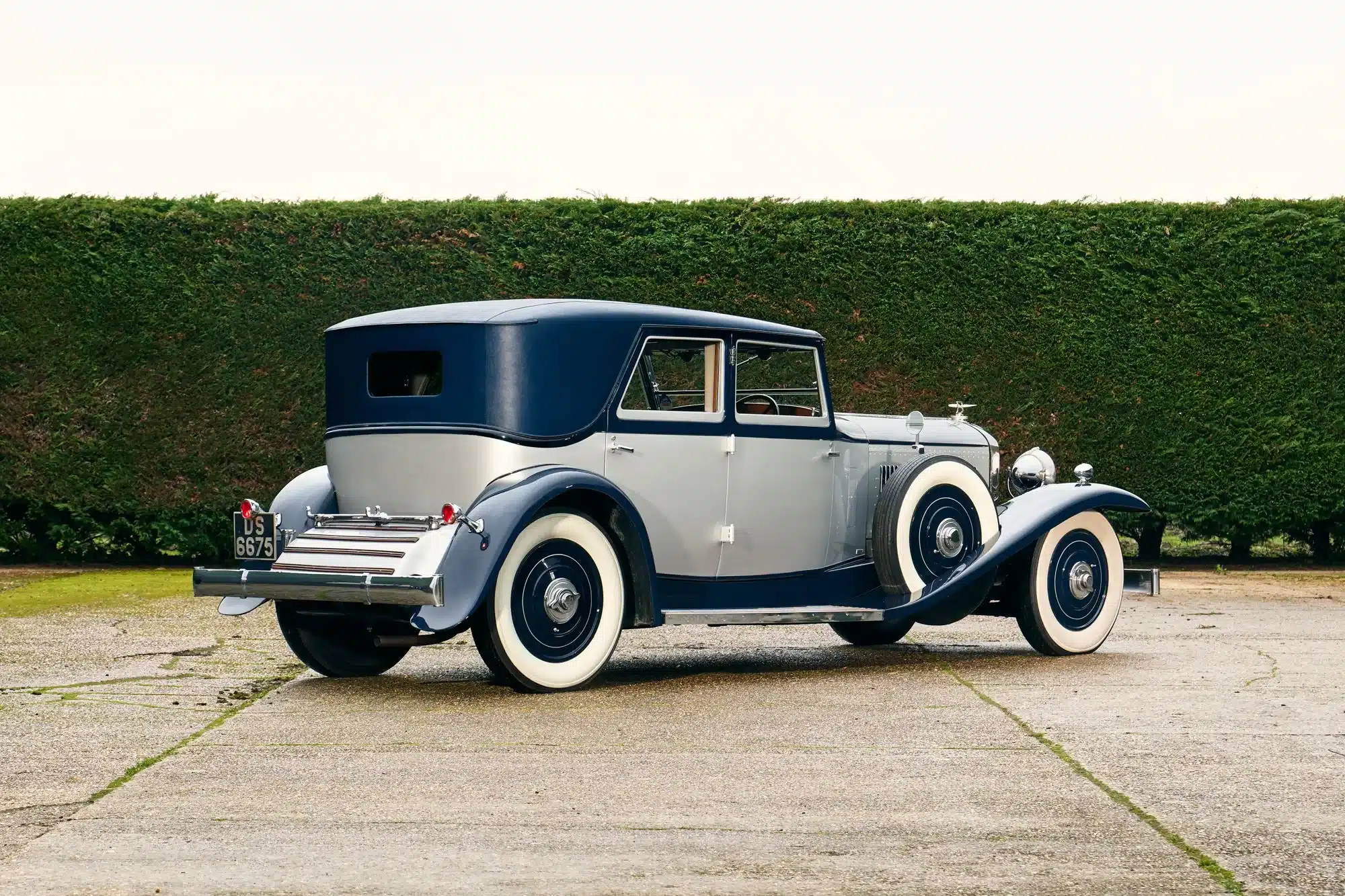 Seitenansicht eines restaurierten 1930 Bentley Speed Six mit zweifarbiger Schutte-Karosserie und Weißwandreifen auf einem Hof vor einer grünen Hecke.