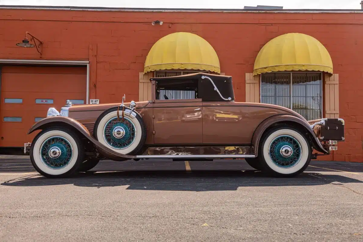 Seitenansicht eines braunen 1930 Stutz MB Convertible Coupe mit markanten Speichenrädern und Verdeck, vor orangefarbener Wand geparkt.