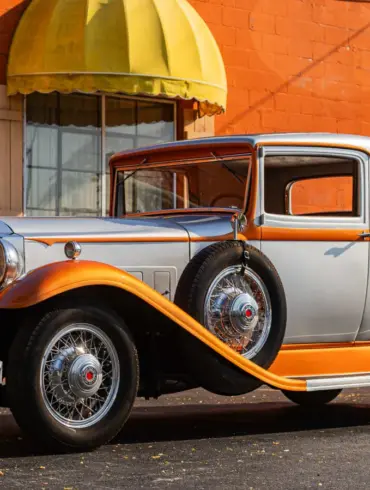 Front- und Seitenansicht eines silber-orangefarbenen Packard Model 902 Eight Coupé von 1932, geparkt vor einem Gebäude mit gelben Markisen.
