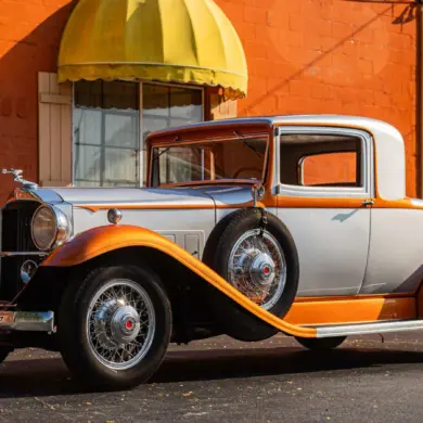 Front- und Seitenansicht eines silber-orangefarbenen Packard Model 902 Eight Coupé von 1932, geparkt vor einem Gebäude mit gelben Markisen.