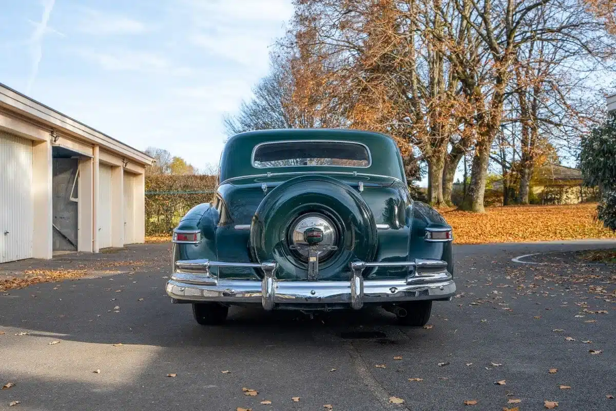 Heckansicht eines dunkelgrünen Lincoln Continental Coupé aus dem Jahr 1948, geparkt auf einem asphaltierten Platz vor Garagen und herbstlichen Bäumen.