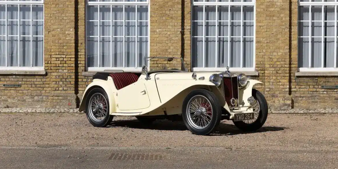 Seitliche Ansicht eines restaurierten 1948 MG TC Midget in Beige mit roten Ledersitzen, geparkt vor einem Gebäude mit gelber Backsteinfassade und großen Fenstern.