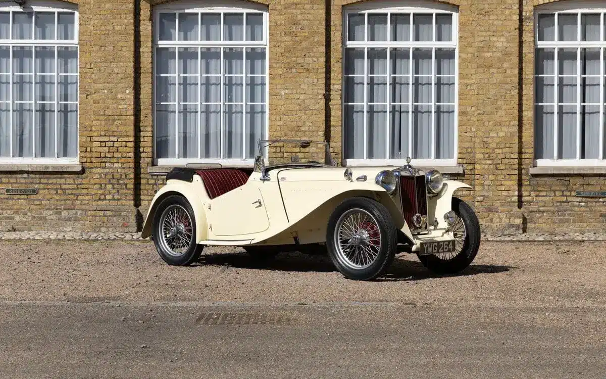 Seitliche Ansicht eines restaurierten 1948 MG TC Midget in Beige mit roten Ledersitzen, geparkt vor einem Gebäude mit gelber Backsteinfassade und großen Fenstern.