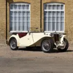 Seitliche Ansicht eines restaurierten 1948 MG TC Midget in Beige mit roten Ledersitzen, geparkt vor einem Gebäude mit gelber Backsteinfassade und großen Fenstern.