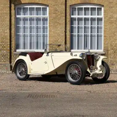 Seitliche Ansicht eines restaurierten 1948 MG TC Midget in Beige mit roten Ledersitzen, geparkt vor einem Gebäude mit gelber Backsteinfassade und großen Fenstern.