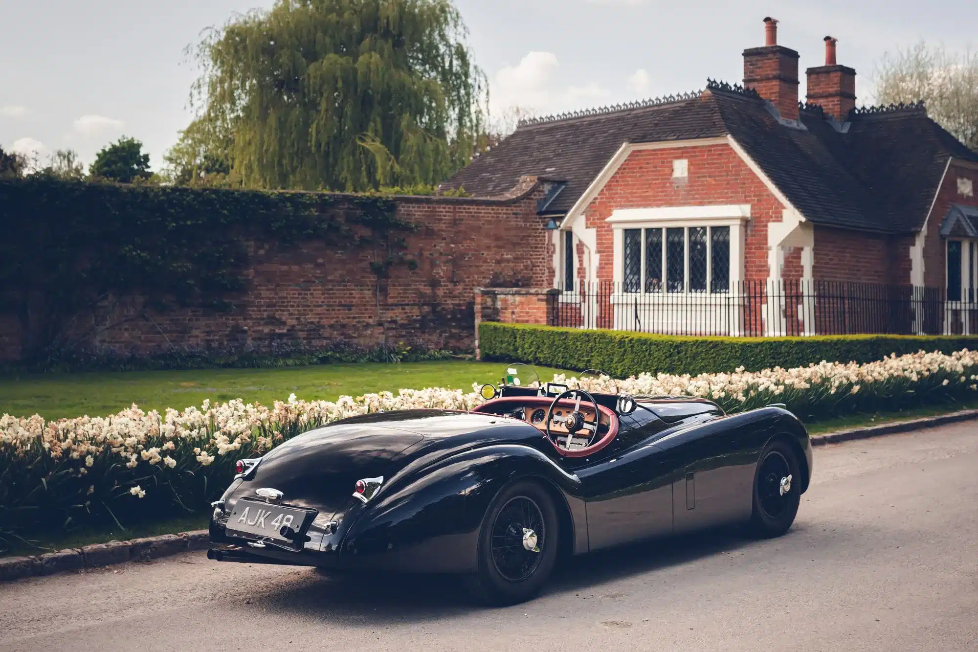 Schwarzer Jaguar XK120 Roadster von 1951 mit rotem Armaturenbrett, geparkt vor einem Backsteinhaus und Blumenbeeten.