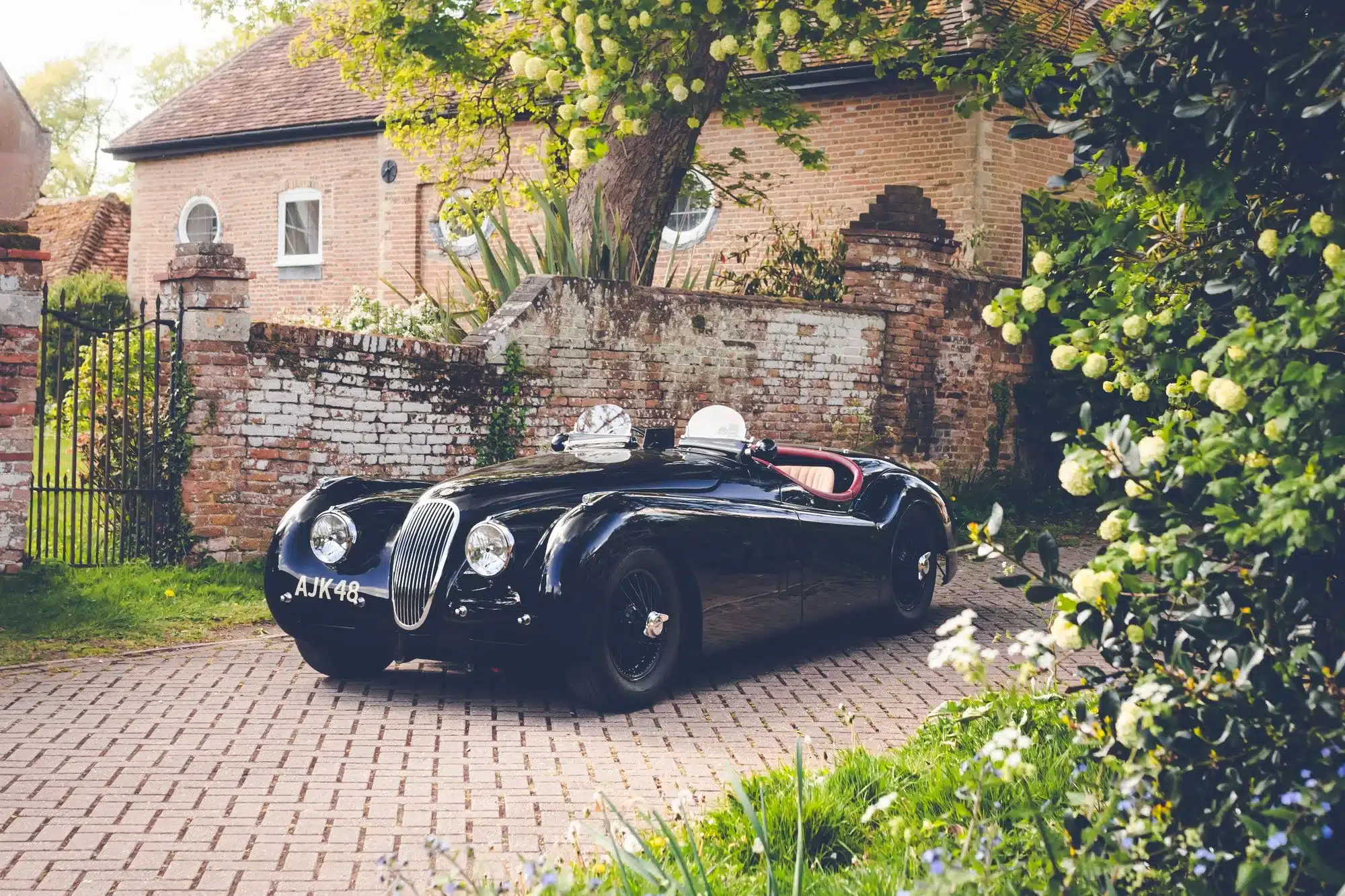 Schwarzer Jaguar XK120 Competition Roadster von 1951 mit Biscuit-Rot-Innenraum, geparkt auf einem Pflasterweg vor einer Backsteinmauer und einem Baum