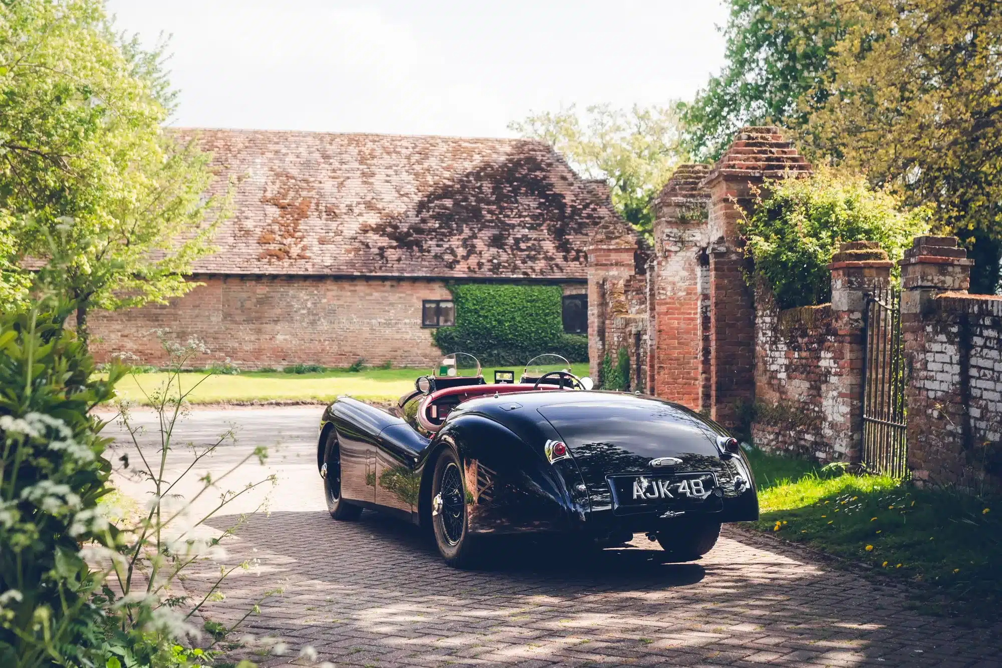 Schwarzer Jaguar XK120 Competition Roadster von 1951 mit rotem und beigem Innenraum vor alten Backsteinmauern und Hofeinfahrt von hinten fotografiert.