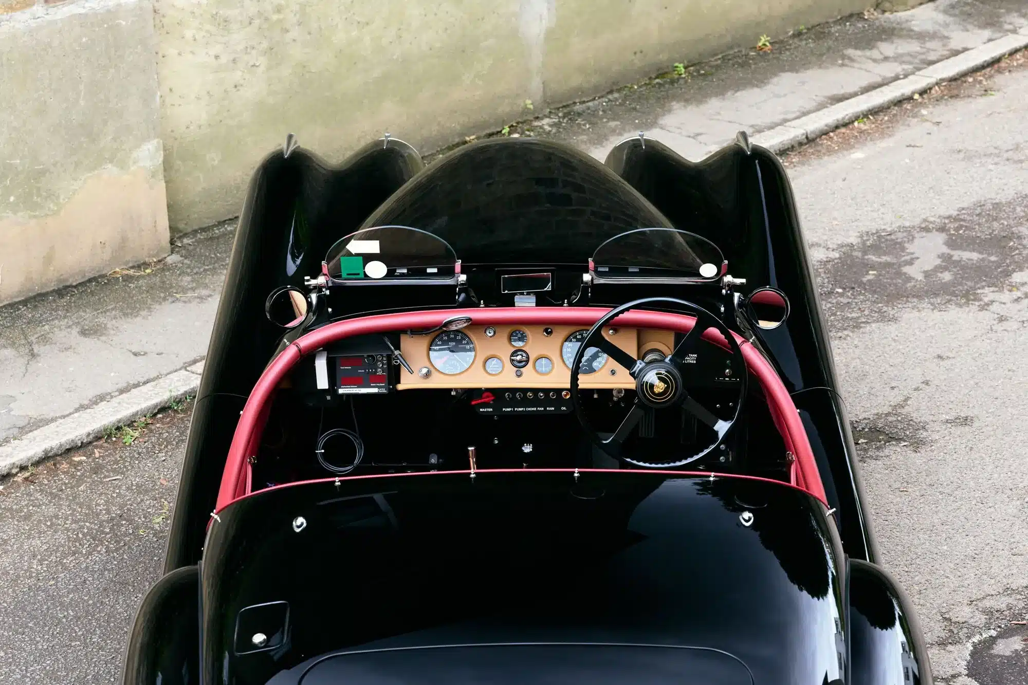 Blick von oben auf das Cockpit eines schwarzen Jaguar XK120 Roadster (Baujahr 1951) mit rotem Überrollbügel und analoger Instrumententafel.