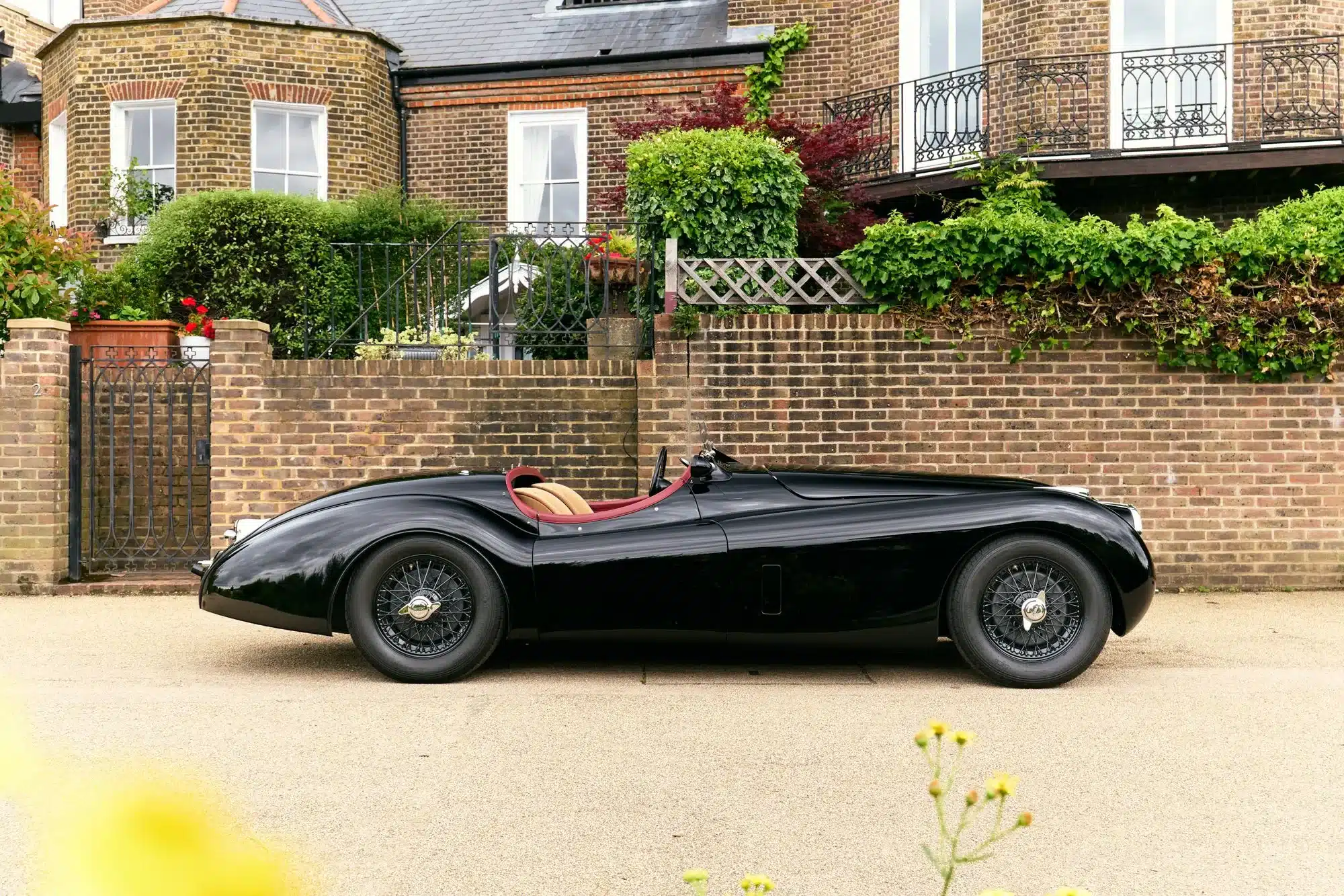 Schwarzer Jaguar XK120 Competition Roadster von 1951 mit Biscuit-Rotem Interieur vor einer Backsteinmauer abgebildet.