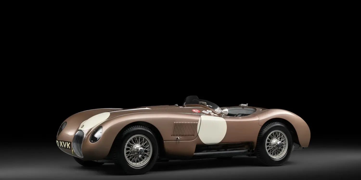 Seitenansicht des Jaguar C-Type XKC 018 in Bronze mit weißen Startnummernfeldern vor schwarzem Hintergrund