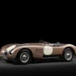 Seitenansicht des Jaguar C-Type XKC 018 in Bronze mit weißen Startnummernfeldern vor schwarzem Hintergrund