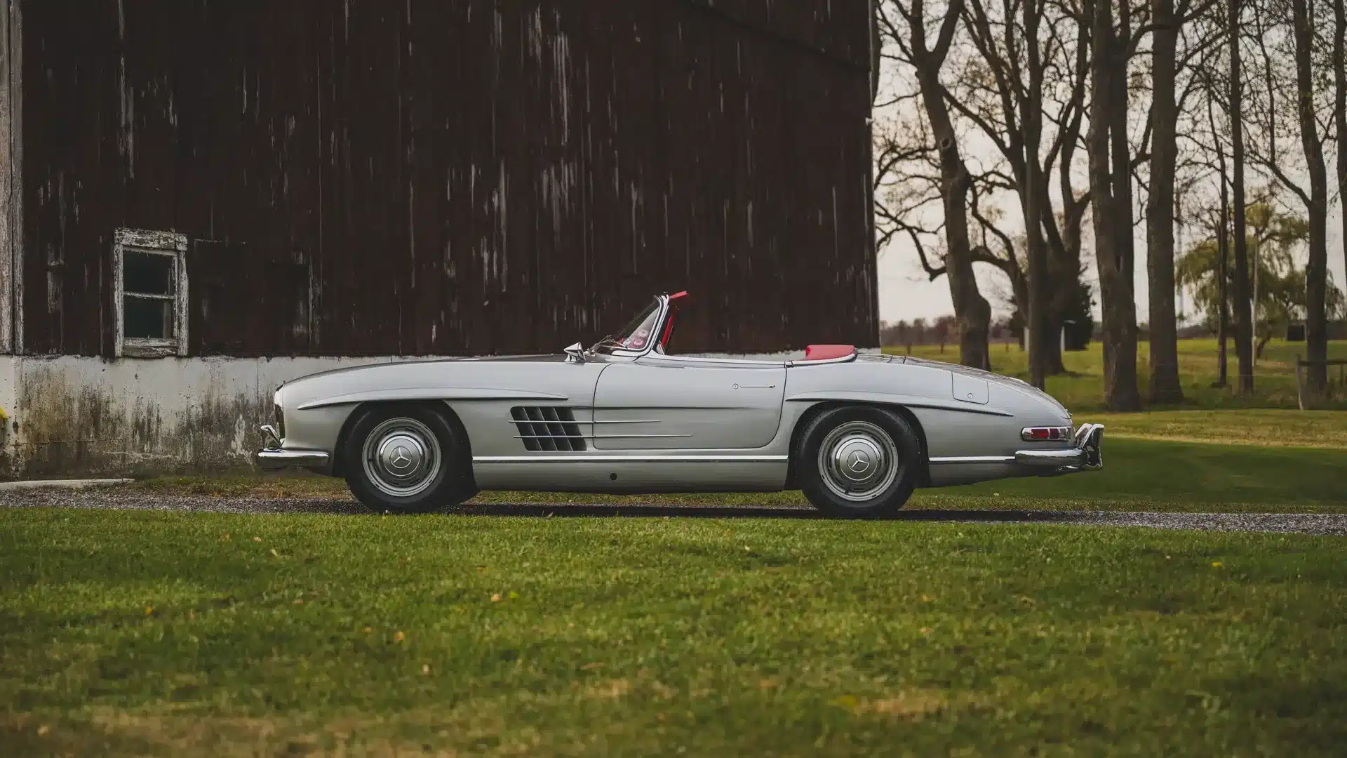 Mercedes-Benz 300 SL Roadster, Baujahr 1957, silber mit rotem Dach und rotem Lederinterieur, seitlich fotografiert vor einer Scheune.