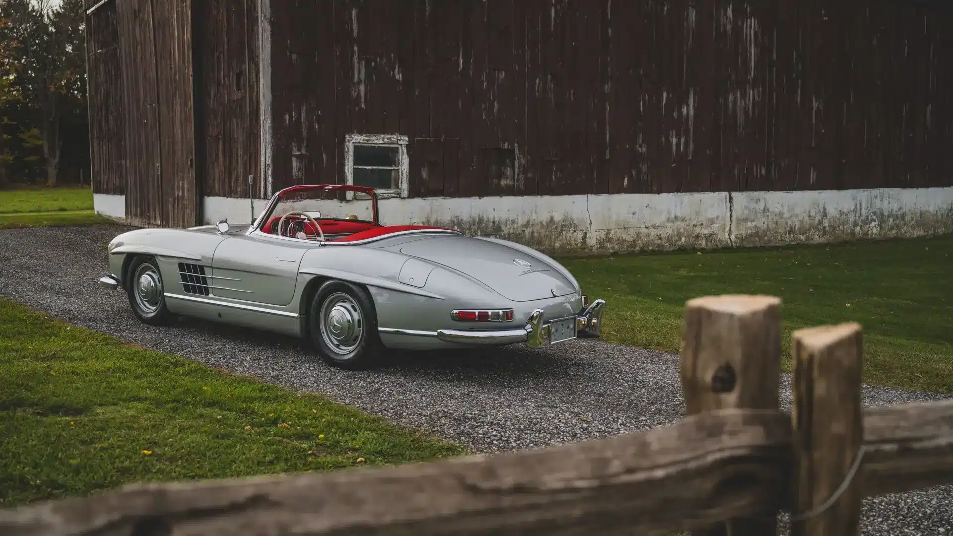 Silberner Mercedes-Benz 300 SL Roadster von 1957 mit rotem Verdeck, seitlich geparkt vor einer alten Holzscheune.