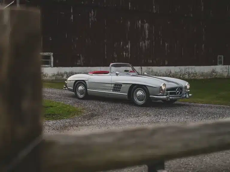 Mercedes-Benz 300 SL Roadster von 1957 in Silber mit rotem Lederinterieur vor einer Scheune auf einem Schotterweg.