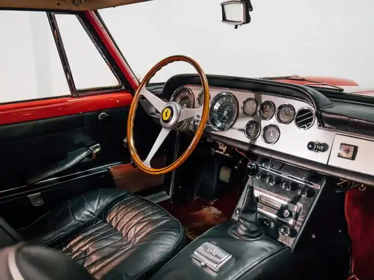 Innenraum eines Ferrari 250 GTE Series III mit schwarzem Leder und Holzlenkrad, Blick auf Armaturenbrett und Schaltknauf.