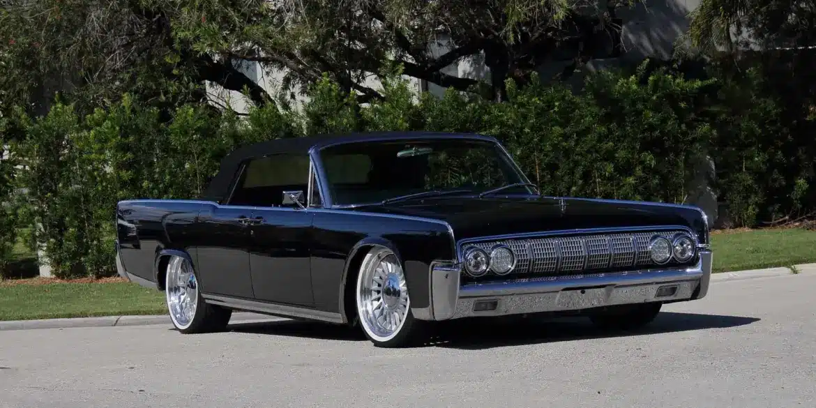 Lincoln Continental Cabriolet von 1964 mit schwarzen Lack und geöffnetem Verdeck, modernisiert mit großen Felgen, stehend auf einer Straße vor grüner Hecke.