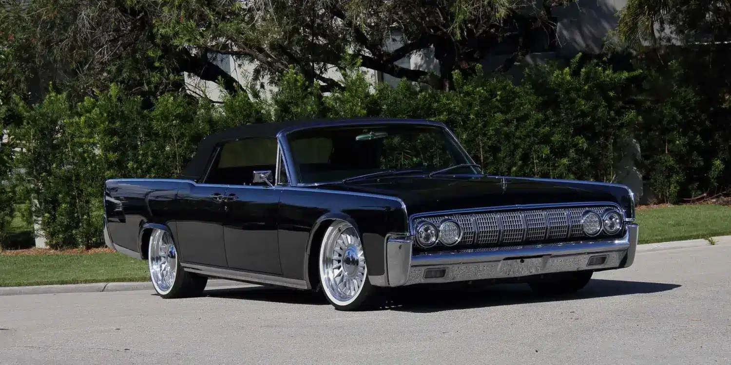 Lincoln Continental Cabriolet von 1964 mit schwarzen Lack und geöffnetem Verdeck, modernisiert mit großen Felgen, stehend auf einer Straße vor grüner Hecke.