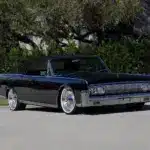 Lincoln Continental Cabriolet von 1964 mit schwarzen Lack und geöffnetem Verdeck, modernisiert mit großen Felgen, stehend auf einer Straße vor grüner Hecke.