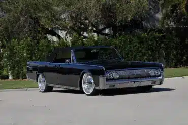 Lincoln Continental Cabriolet von 1964 mit schwarzen Lack und geöffnetem Verdeck, modernisiert mit großen Felgen, stehend auf einer Straße vor grüner Hecke.