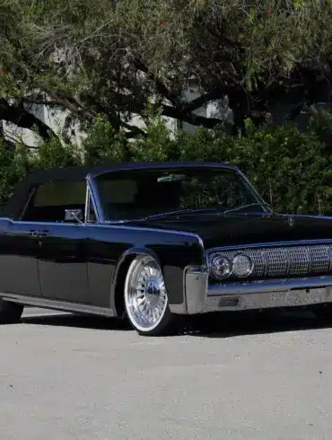 Lincoln Continental Cabriolet von 1964 mit schwarzen Lack und geöffnetem Verdeck, modernisiert mit großen Felgen, stehend auf einer Straße vor grüner Hecke.