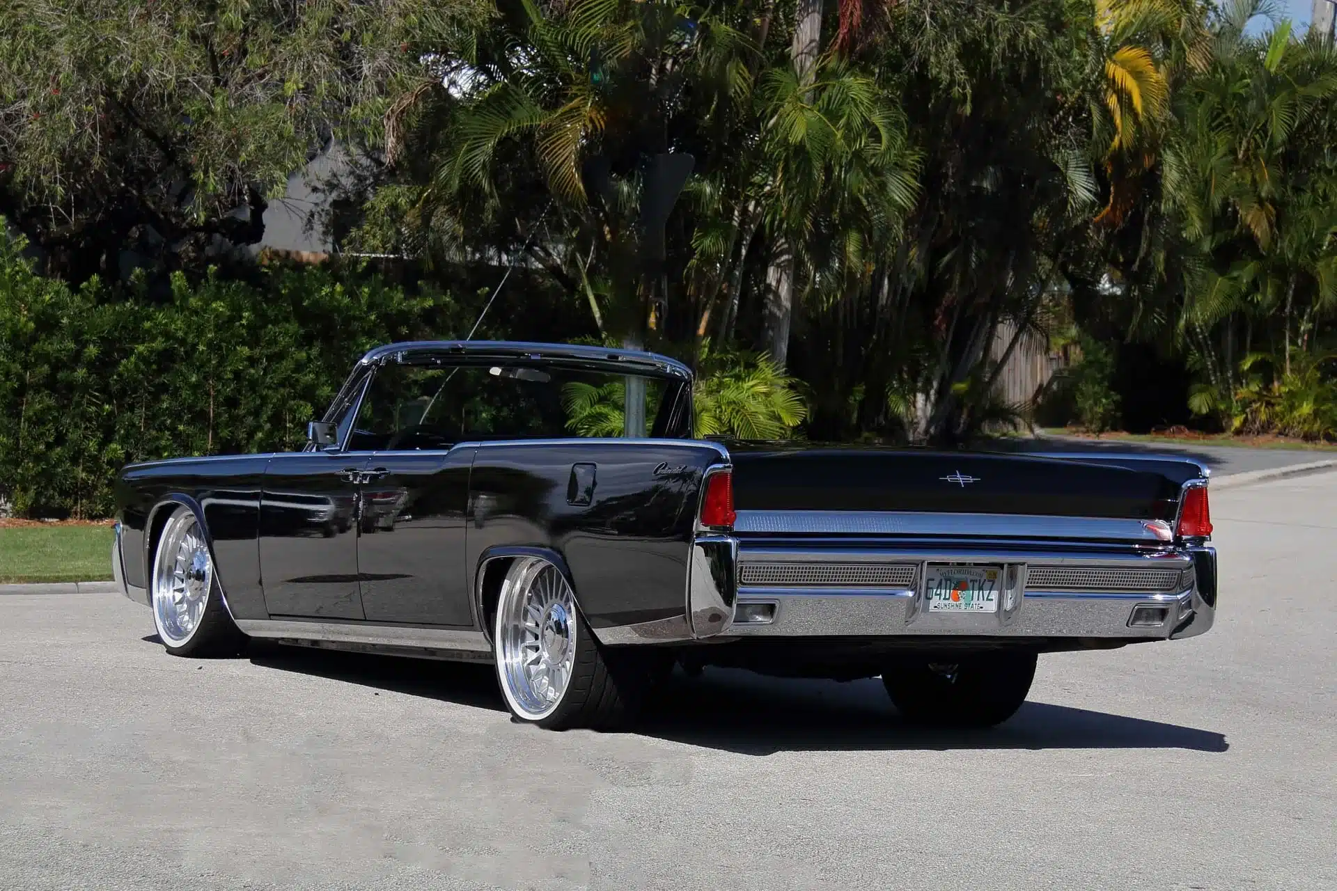 Seiten- und Heckansicht eines schwarzen 1964 Lincoln Continental Cabriolets mit geöffnetem Verdeck, großen Chromfelgen und charakteristischen hinten angeschlagenen Türen, geparkt vor Palmen.