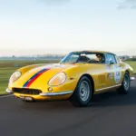 Gelber Ferrari 275 GTB Long Nose Alloy Baujahr 1965 mit blauen und roten Streifen fährt auf einer Rennstrecke.