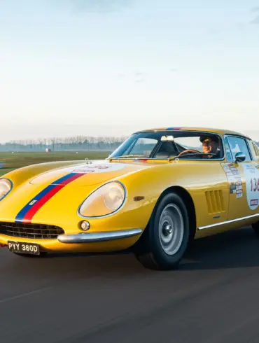Gelber Ferrari 275 GTB Long Nose Alloy Baujahr 1965 mit blauen und roten Streifen fährt auf einer Rennstrecke.