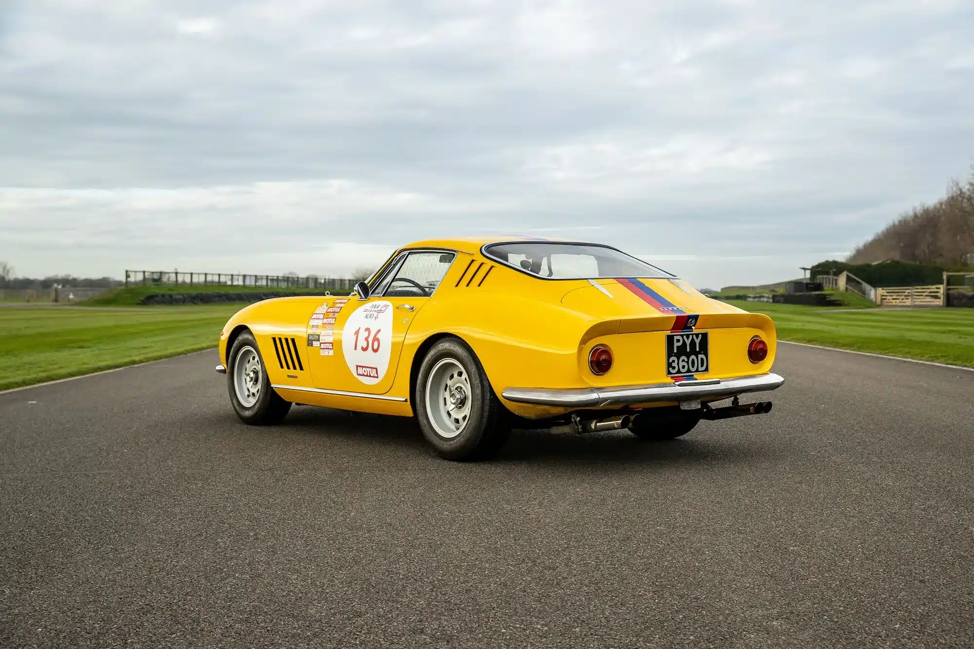 Gelber Ferrari 275 GTB Long Nose Alloy von 1965 mit Startnummer 136 und Rallyestreifen auf einer Landstraße.
