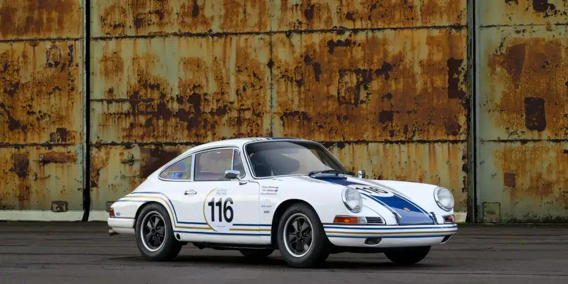 Historischer Porsche 911 in Rennausführung, Baujahr 1965, weiß mit blauen Rennstreifen und Startnummer 116, vor einer rostigen Wand.