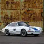 Historischer Porsche 911 in Rennausführung, Baujahr 1965, weiß mit blauen Rennstreifen und Startnummer 116, vor einer rostigen Wand.