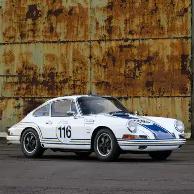 Historischer Porsche 911 in Rennausführung, Baujahr 1965, weiß mit blauen Rennstreifen und Startnummer 116, vor einer rostigen Wand.