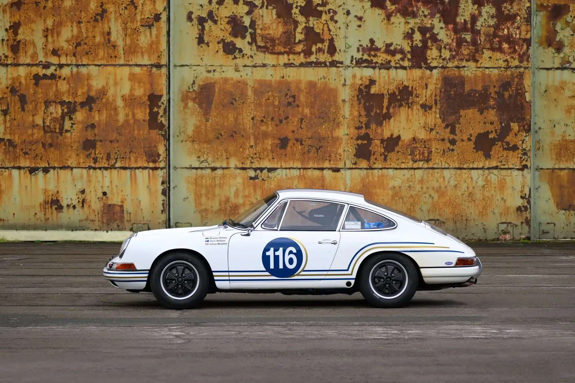 Seitenansicht eines weißen Porsche 911 SWB von 1965 mit Startnummer 116 und Rennsportumbau, geparkt vor einer rostigen Metallwand