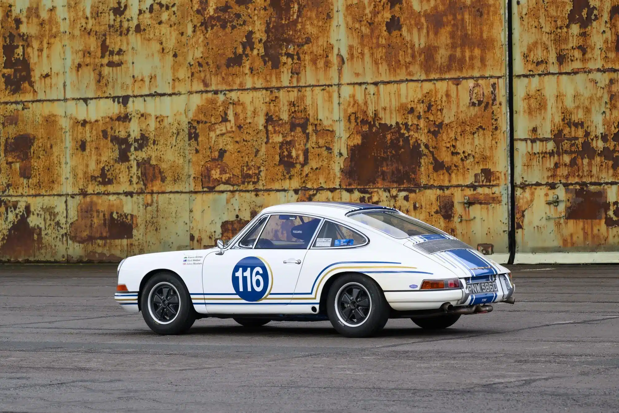 Seitenansicht eines weißen Porsche 911 SWB, Baujahr 1965, als historischer Rennwagen mit Startnummer 116 vor einer rostigen Wand.