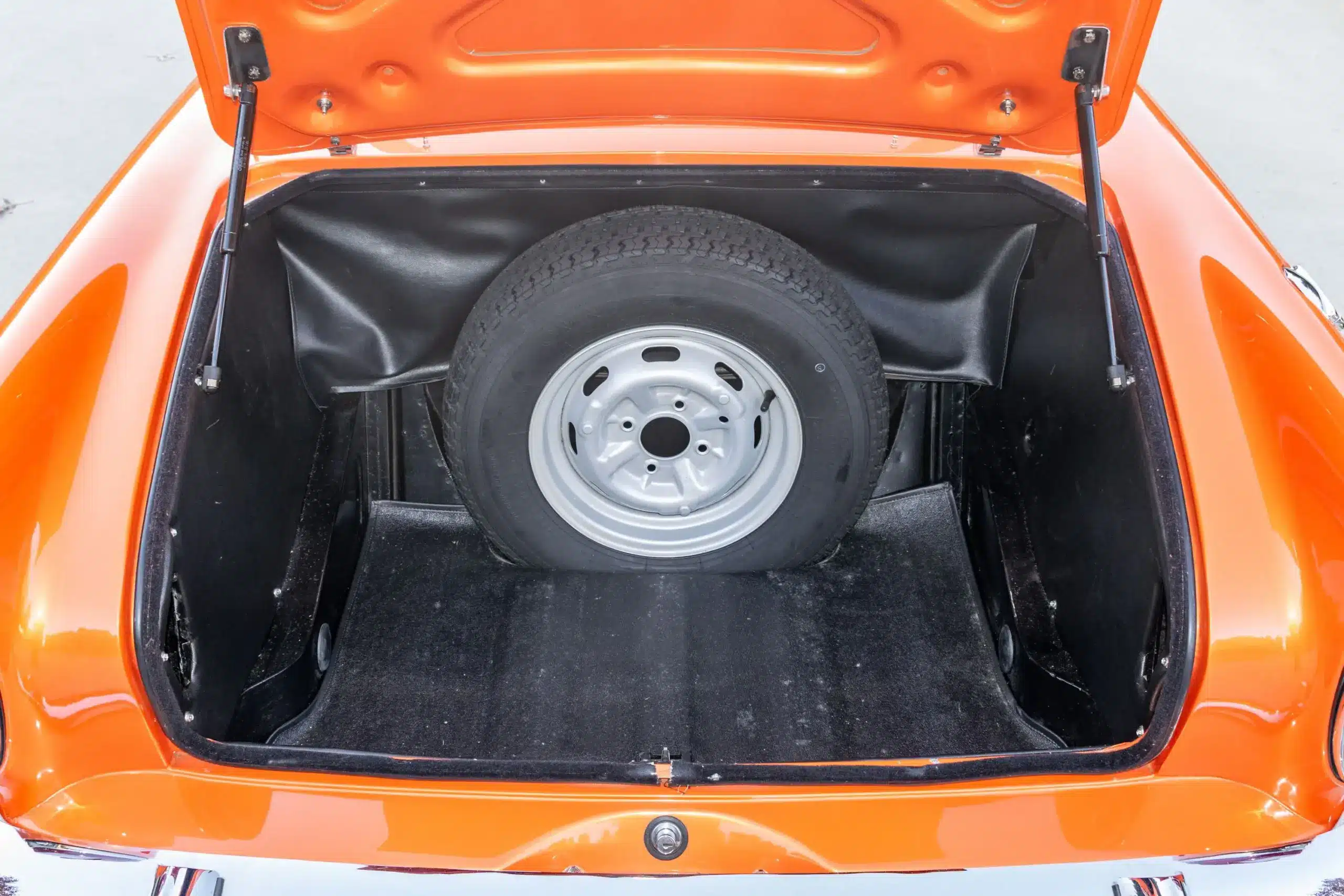 Geöffneter Kofferraum eines Sunbeam Tiger Mk1, Reserverad vor schwarzer Innenauskleidung, orangefarbene Lackierung sichtbar.