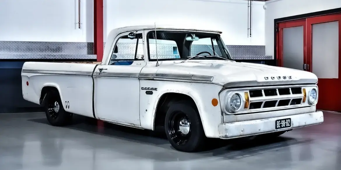 Seitenansicht eines weißen Dodge D100 Pickups von 1968 mit originaler Lackierung und altersbedingter Patina, geparkt in einer hellen Halle.