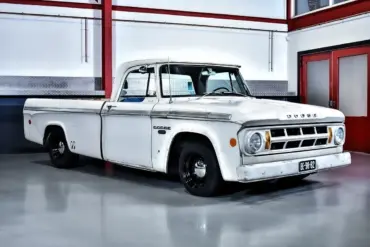 Seitenansicht eines weißen Dodge D100 Pickups von 1968 mit originaler Lackierung und altersbedingter Patina, geparkt in einer hellen Halle.