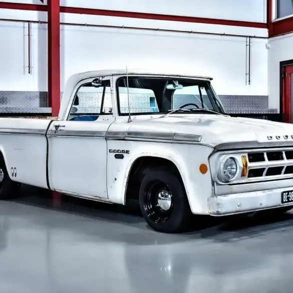 Seitenansicht eines weißen Dodge D100 Pickups von 1968 mit originaler Lackierung und altersbedingter Patina, geparkt in einer hellen Halle.