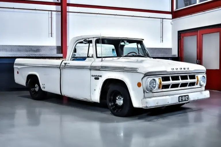 Seitenansicht eines weißen Dodge D100 Pickups von 1968 mit originaler Lackierung und altersbedingter Patina, geparkt in einer hellen Halle.