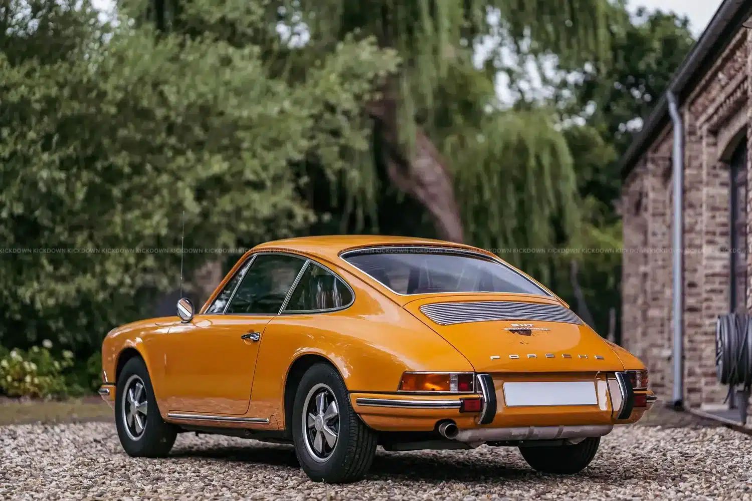 Heckansicht eines orangefarbenen Porsche 911 T Sportomatic von 1969, stehend auf einem Kiesweg neben einem Gebäude.