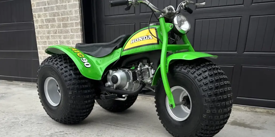 Grünes dreirädriges Honda ATC 90 Geländefahrzeug aus dem Jahr 1972, stehend auf Beton vor schwarzem Garagentor