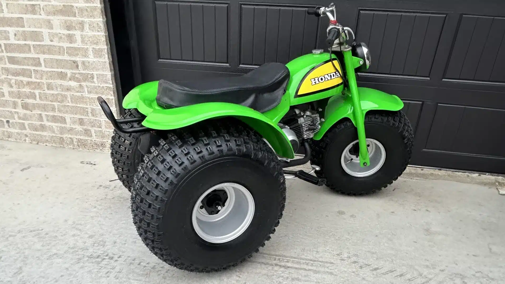 Seltener, grüner Honda ATC 90 Dreirad-Geländewagen Baujahr 1972 mit Ballonreifen und puristischem Retro-Design, abgestellt vor einer Garage.
